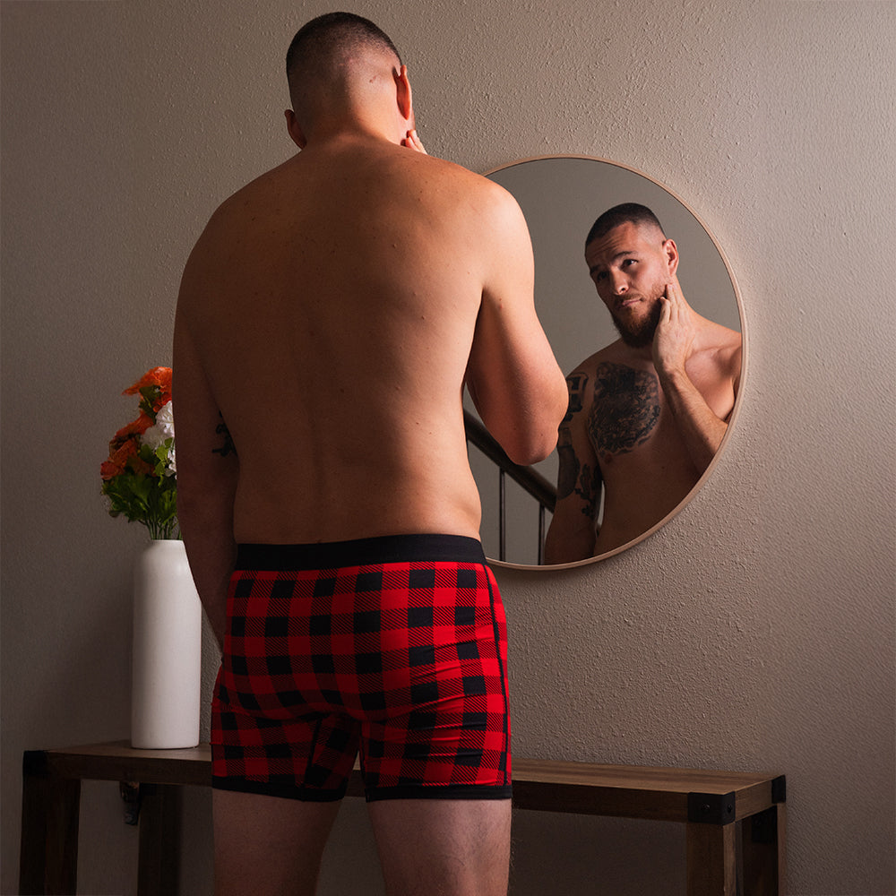 Man in red and black holiday plaid dual pouch boxer briefs looking at himself in a mirror.