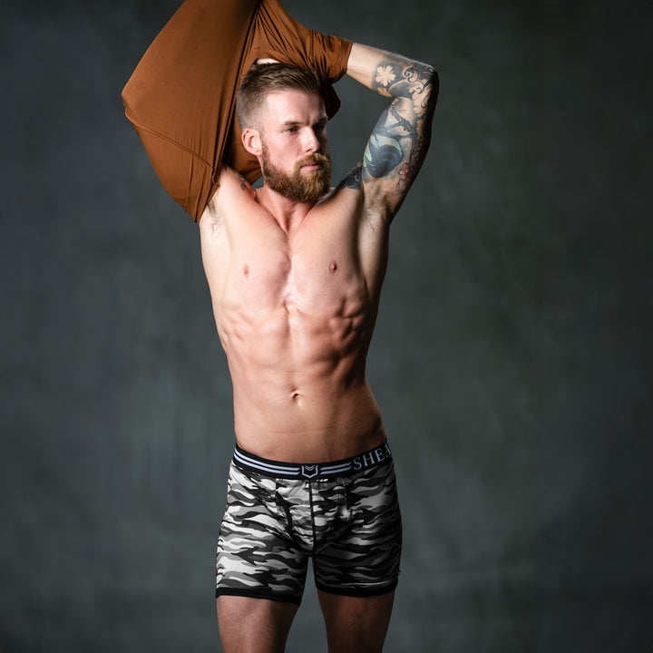 Male model in front of studio backdrop wearing SHEATH polyester boxer briefs in winter gray camo pattern featuring dual pouch ergonomic support, breathable moisture-wicking fabric, anti-chafing design, and a branded elastic waistband with black and white stripes right angle view