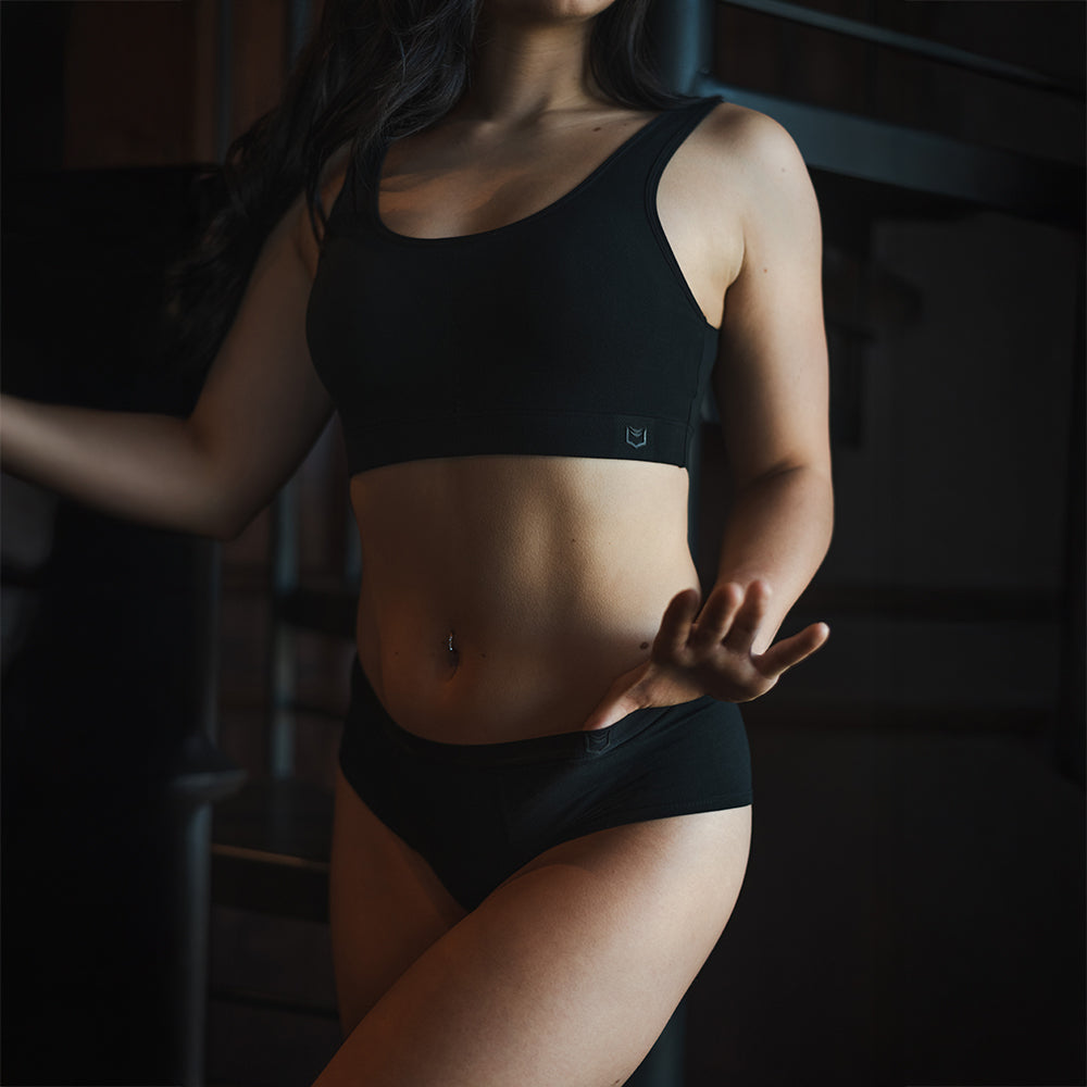 Front view of a female model wearing SHEATH Black padded sports bralette and matching cheeky boyshorts made with breathable bamboo mesh fabric, supportive ergonomic design, and a subtle branded band for comfortable athletic wear