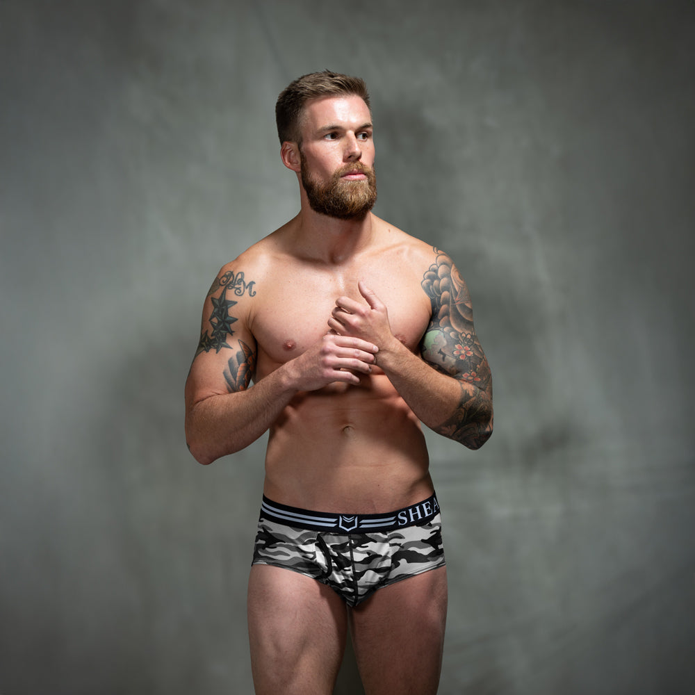 Male model in front of studio backdrop wearing SHEATH camo American briefs in black, gray, and white tones with dual pouch ergonomic support, breathable moisture-wicking polyester fabric, anti-chafing design, and branded elastic waistband. front view