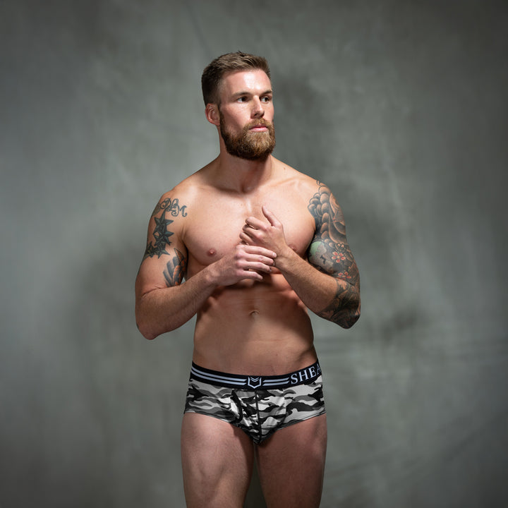 Male model in front of studio backdrop wearing SHEATH camo American briefs in black, gray, and white tones with dual pouch ergonomic support, breathable moisture-wicking polyester fabric, anti-chafing design, and branded elastic waistband. front view
