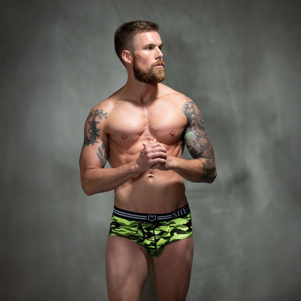 Male model in front of studio backdrop wearing SHEATH American briefs in green camo pattern featuring dual pouch support, breathable polyester fabric, ergonomic fit, anti-chafing design, and a branded elastic waistband. front view