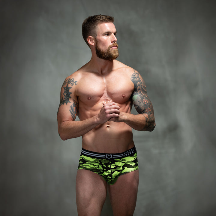 Male model in front of studio backdrop wearing SHEATH American briefs in green camo pattern featuring dual pouch support, breathable polyester fabric, ergonomic fit, anti-chafing design, and a branded elastic waistband. front view