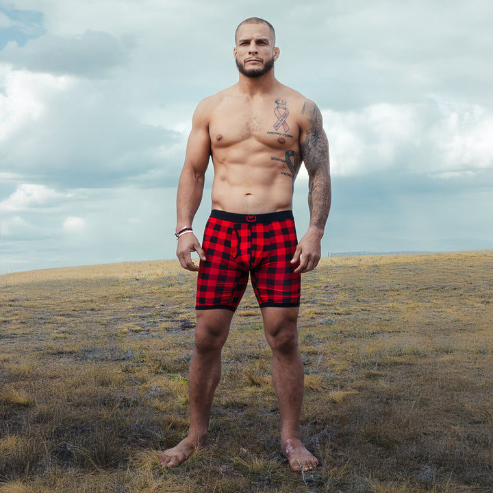 Man wearing red and black holiday plaid dual pouch boxer briefs standing in a grassy field with a cloudy sky.