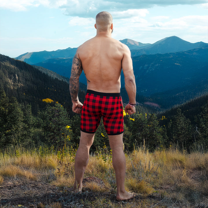 Man wearing red and black holiday plaid dual pouch boxer briefs standing on a mountain trail with mountains in the background