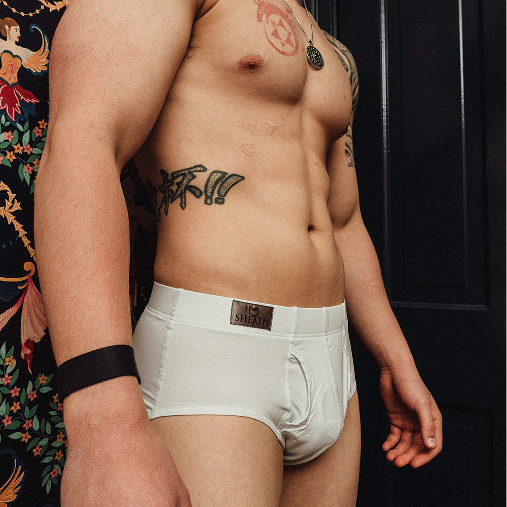 Man wearing white boxer briefs with a brand logo, standing against a dark background. Male model wearing SHEATH Black men's brief with dual pouch ergonomic support, made from breathable modal fabric featuring moisture-wicking and anti-chafing properties for all-day comfort.#color_black