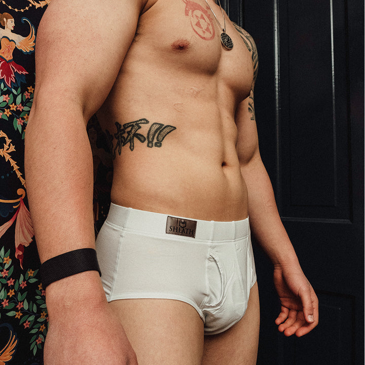Man wearing white boxer briefs with a brand logo, standing against a dark background. Male model wearing SHEATH Black men's brief with dual pouch ergonomic support, made from breathable modal fabric featuring moisture-wicking and anti-chafing properties for all-day comfort.#color_black